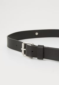 Cinturón de cuero negro con una textura suave, que presenta una hebilla de metal plateado y un lazo seguro. Múltiples agujeros de ajuste para el tamaño.