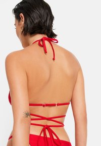 Rode bikini top met halternekstrikjes, voorzien van een textuurafwerking en meerdere gekruiste banden op de rug, gezekerd met ronde hardware.