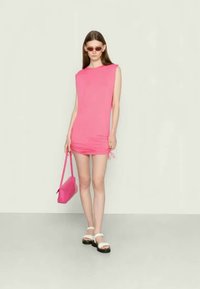 Vestido ajustado rosa em jersey com mangas curtas, laterais franzidas, combinado com sandálias brancas e uma bolsa rosa. Textura suave e design simples.