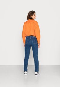Camisa laranja de botão, com corte solto e pregas nas costas. Jeans skinny azuis, lavagem média com dois bolsos traseiros. Ténis brancos com detalhes pretos.