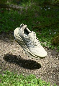 Chaussure de randonnée beige avec une tige en mesh, un branding prominent, une surface texturée et une semelle robuste, surplombant un fond de gravier et d'herbe.