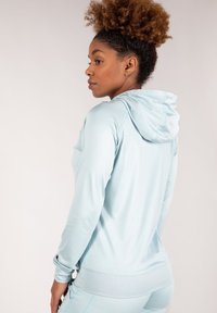 Haut de sport bleu clair avec capuche à cordon, manches longues, texture lisse et coutures contrastées, associé à un bas de sport assorti.