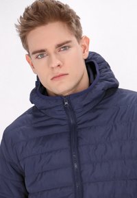 Marine blauwe pufferjack met een capuchon, voorzien van een gewatteerd ontwerp, ritssluiting en een gladde, glanzende textuur. Ideaal voor buitenkleding.