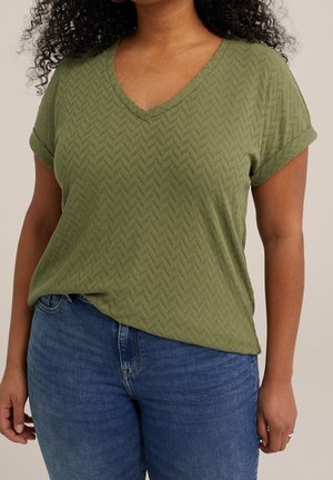 Vrouw draagt een groene, kortebovenmouwen, gestructureerde V-hals top, ingestopt in een blauwe spijkerbroek, staand tegen een effen achtergrond.