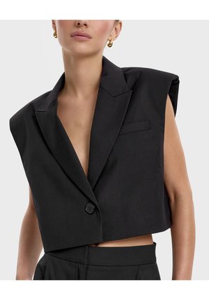 Kobieta ubrana w czarną, bezrękawną, krótką marynarkę z poduszonymi ramionami, zapinaną na pojedynczy guzik, oraz złote kolczyki-koła, na tle jasnego, jednolitego tła.