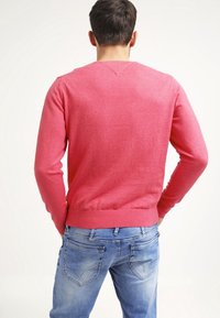 Pullover rosso lavorato a maglia con maniche lunghe, vita aderente e una texture liscia, abbinato a jeans azzurri chiari con dettagli di cucitura sottili.