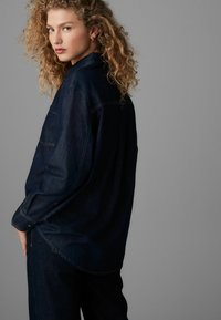 Dunkelblauses Denimhemd mit langen Ärmeln, Knopfmanschetten, Kragen, einer Brusttasche und leicht geschwunghem Saum. Getragen mit passender Jeans.