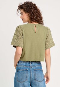 Top court vert olive avec des manches courtes en dentelle texturée et un détail de dos en forme de clé, associé à un jean en denim bleu taille haute.