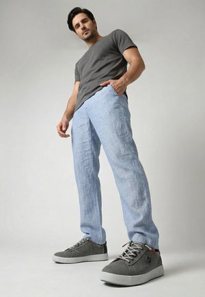 Mann steht mit einer Hand in der hellblauen Hosentasche, trägt ein dunkelgraues T-Shirt und graue Sneaker vor einem einfarbigen Hintergrund.