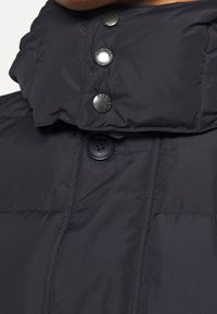 Mörkblå puffjacka med hög krage, med metallknappar och en strukturerad tygkonstruktion. Sömnadsdetaljer framhäver designen.