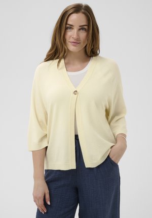 Vrouw met lang bruin haar draagt een lichtgele cardigan met één knoop, een witte top en marineblauwe broek met trekkoord, staand tegen een neutrale achtergrond.