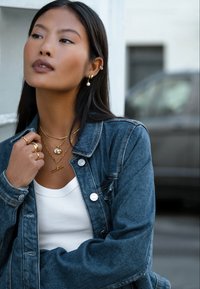 Τζιν μπουφάν με μεταλλικά κουμπιά, λευκό αμάνικο τοπ, στρωμένα χρυσά κολιέ και κρίκοι με λεπτομέρεια πέρλας. Μακριά σκούρα μαλλιά.