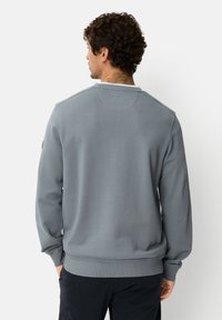 Grijze trui met geribde manchetten en zoom, ronde hals, gladde textuur en een patch-detail op de linker mouw. Achteraanzicht getoond.