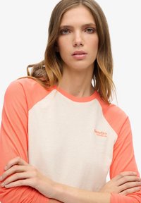 Young woman with long light brown hair wearing a white and coral raglan sleeve shirt with folded arms against a white background.