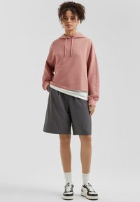 Lichtroze geïmmersede hoodie met lange mouwen, gecombineerd met donkergrijze losse shorts; model draagt witte sokken en zwarte sneakers.