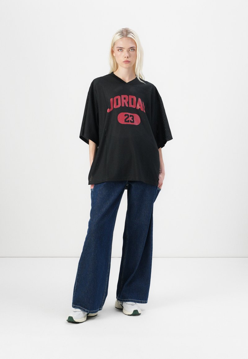 T-shirt oversized en maille noire avec un graphique rouge "JORDAN 23", associé à un jean bleu large et à des baskets blanches avec des accents verts.