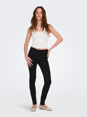 Młoda kobieta z długimi falującymi włosami, ubrana w biały bezrękawnik, czarne wąskie spodnie i beżowe baleriny, stoi z jedną ręką na biodrze.