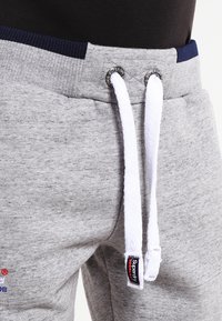 Graue Jogginghose mit strukturierter Oberfläche, weißen Kordeln, marineblauen Akzenten am Bund und einem Superdry-Logo an der Seite.