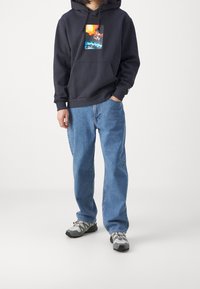 Person wearing a black hoodie with a colorful graphic, light blue jeans, and gray hiking shoes, standing with one hand in pocket.