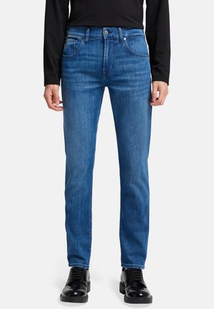 Jean en denim bleu avec une coupe droite, doté d'un design à cinq poches, de passants de ceinture et d'un léger délavage. Associé à des chaussures noires.