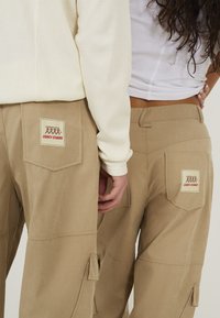 Beige cargobroeken hebben een patch pocket met een "Legacy Studios" label, textielstructuur en zichtbare stikseldetails.