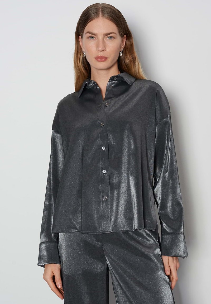 Camicia lunghe maniche grigia metallica con colletto a punta, caratterizzata da una texture liscia e una vestibilità comoda. Bottoni visibili sul davanti.