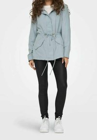 Veste à capuche bleu clair avec taille cintrée, deux poches latérales et cordon de serrage. Associée à des leggings noirs et des baskets blanches.