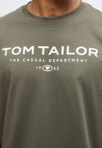 Λαδί πράσινη μπλούζα με λευκό κείμενο "TOM TAILOR THE CASUAL DEPARTMENT 1962" στο κέντρο του στήθους.