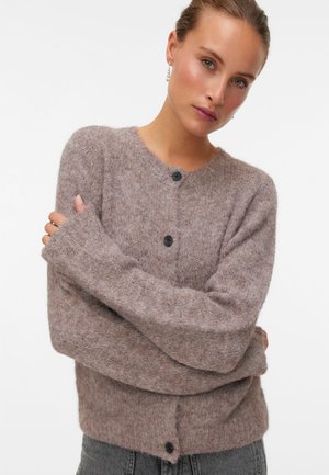 Jeune femme aux cheveux châtain clair portant un cardigan marron doux boutonné et un jean gris foncé, les bras croisés sur un fond uni.