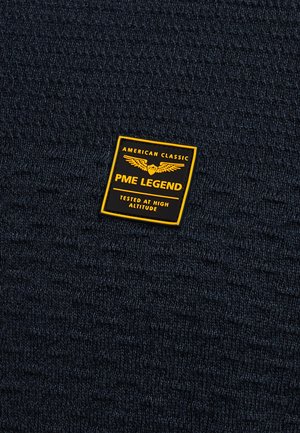 Tmavomodrá pletená látka s textúrovaným vzorom, ktorú zdobí žlto-čierna obdĺžniková nálepka s nápismi "PME LEGEND" a "TESTED AT HIGH ALTITUDE".