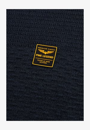 Tmavomodrá pletená látka s textúrovaným vzorom, ktorú zdobí žlto-čierna obdĺžniková nálepka s nápismi "PME LEGEND" a "TESTED AT HIGH ALTITUDE".