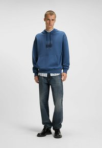 Felpa blu con tasca frontale e logo, indossata sopra una camicia a righe. Abbinata a jeans scuri e sbiaditi e stivali neri stringati.