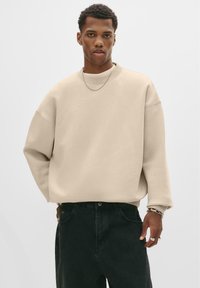 Hanorac oversized bej cu guler rotund, manșete ribbed �și o siluetă relaxată, asociat cu blugi verzi închis și accente de bijuterii din argint.