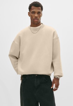 Beige oversized sweatshirt met een ronde hals, geribbelde manchetten en een losse pasvorm, gecombineerd met donkergroene jeans en zilverkleurige sieraden.