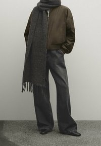 Brauner Bomberjacke mit Reißverschluss, grauer Schal mit Fransen, dunkle weite Jeans und schwarze spitz zulaufende Schuhe vor einem neutralen Hintergrund.