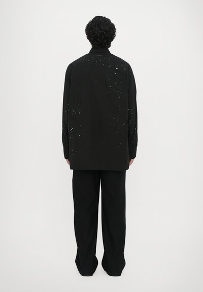 Chemise noire à manches longues avec une coupe ample, arborant des motifs de éclaboussures de peinture sur le dos, associée à un pantalon noir à jambes larges.