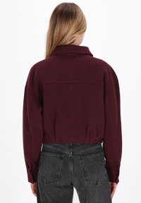 Burgunderfarbene verkürzte Jacke mit kragen, langen Ärmeln und strukturiertem Stoff. Kombiniert mit dunkelgrauen, hochgeschnittenen Jeans, die Rückendetails zeigen.