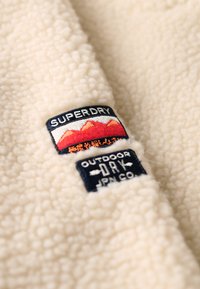 Beżowy polar o teksturowanej powierzchni. Posiada dwa haftowane naszywki: "SUPERDRY" oraz "OUTDOOR DRY JPN CO." w kontrastowych kolorach.