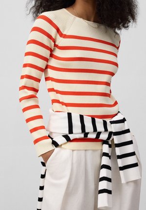 Personne portant un pull crème à rayures rouges horizontales et un pantalon blanc, avec un pull blanc à rayures noires noué autour de la taille.