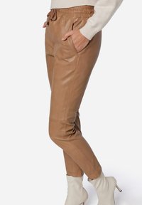 Brune læder joggers med elastisk talje, sidelommer og strømlinet pasform, parret med cremede ankelstøvler, mod en ensfarvet baggrund.