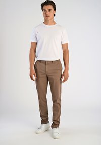 Giovane uomo in piedi, indossando una maglietta bianca a maniche corte, pantaloni marroni e sneakers bianche, su uno sfondo chiaro e uniforme.