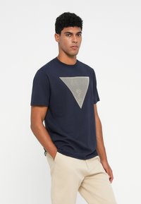 Camiseta de algodón azul marino con un diseño geométrico de triángulos en gris claro en la parte frontal. Cuello redondo estándar, mangas cortas, corte relajado.