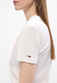 Biała bawełniana koszulka z krótkim rękawem, z pomarańczowym graficznym wzorem oraz małym, haftowanym logo w granatowym, czerwonym i białym kolorze na rękawie.