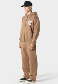 Brauner Zip-Hoodie mit einer Tasche und weißem Grafikdesign, kombiniert mit passenden Hosen. Das Outfit hat eine lockere Passform und besteht aus weichem Stoff. Schwarze Mütze und Sneaker.