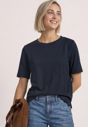 Ung kvinde med kort blondt hår iført en marineblå tekstureret kortærmet skjorte og blå jeans, holder en brun jakke, smilende og kigger mod højre.