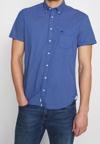 Camicia azzurra a maniche corte con bottoni, con un pattern a griglia, colletto all'italiana e una tasca sul petto. Realizzata in un tessuto leggero.