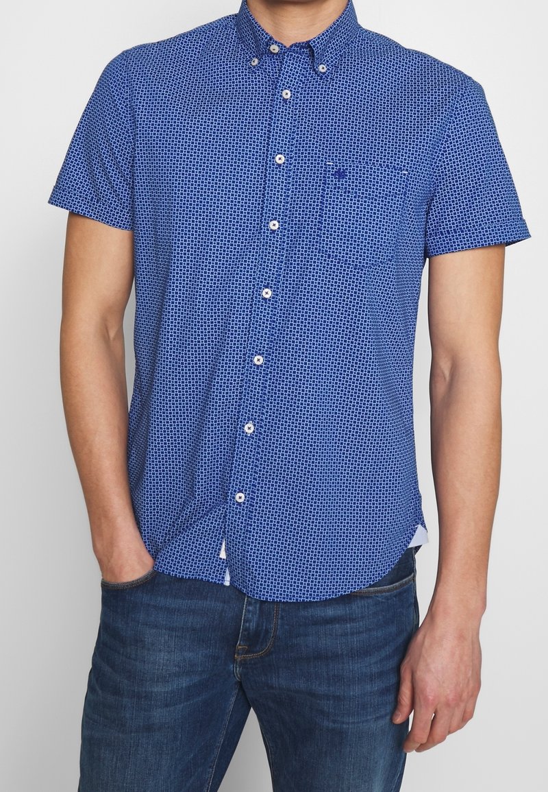 Camicia azzurra a maniche corte con bottoni, con un pattern a griglia, colletto all'italiana e una tasca sul petto. Realizzata in un tessuto leggero.