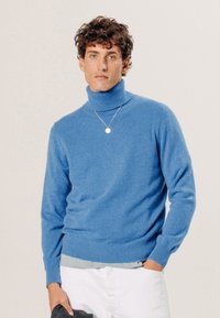 Blauer Rollkragenpullover aus weichem Stoff, mit figurnaher Passform, gerippten Bündchen und Saum, getragen über einem hellgrauen Hemd.