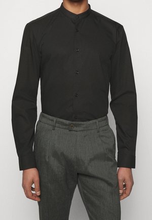 Homme portant une chemise noire cintrée à col officier rentrée dans un pantalon gris taille haute sur mesure, avec fermeture par boutons.