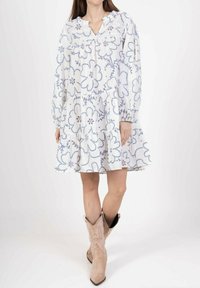 Robe blanche avec broderie florale bleue, manches longues et jupe à volants. Présente un décolleté en V et une coupe décontractée. Des bottes de cowgirl complètent la tenue.